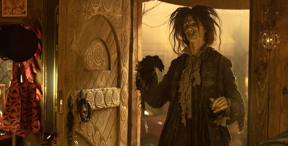 Doug Jones, in character as the zombie Billy Butcherson, stands in a doorway. His hair is messy and his lips are sewn shut, and he is wearing tattered 17th-century style clothing. He is backlight by a yellow light. Assorted merchandise is just inside the door.