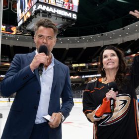 Actor Josh Duhamel holds a microphone in his right hand. He wears a blue suit with a light blue dress shirt. Actor Lauren Graham stands to his left and waves. She wears a black Anaheim Ducks jersey with the gold team logo across the front. They stand on the rink of Honda Center in Anaheim, California.
