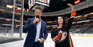 Actor Josh Duhamel holds a microphone in his right hand. He wears a blue suit with a light blue dress shirt. Actor Lauren Graham stands to his left and waves. She wears a black Anaheim Ducks jersey with the gold team logo across the front. They stand on the rink of Honda Center in Anaheim, California.