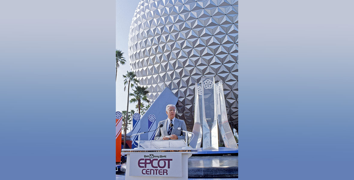 Card Walker, then-Chairman and CEO of The Walt Disney Company (and future Disney Legend), at the lectern at the opening ceremonies for EPCOT on October 1, 1982. He’s wearing a grey suit. Spaceship Earth is seen in the background, as are some palm trees. The former EPCOT Center logo is seen on the lectern.