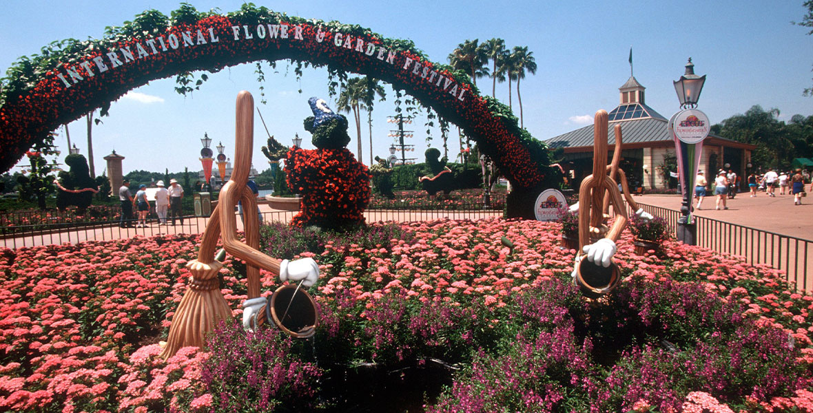 An image from the 1997 EPCOT International Flower & Garden Festival, on a bright sunny day. The name of the festival is seen in white sparkly letters on a flowered arch over a display of light pink flowers, as well as a topiary shaped like Sorcerer Mickey and three broom figures from Fantasia.