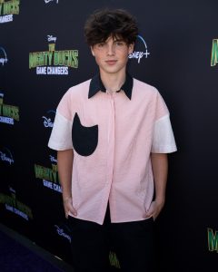 Actor Brady Noon stands on the purple carpet at the premiere event for the Season 2 The Mighty Ducks: Game Changers. He wears a pink and white short sleeved, button down shirt with a black pocket and collar.