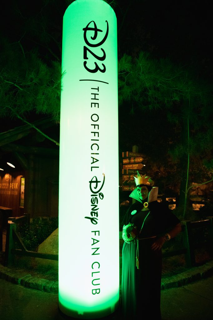A long vertical blow-up tube lit in green with black text reading D23: The Official Disney Fan Club. The guest standing to the right of the glow tube is dressed as the Evil Queen from Snow White and the Seven Dwarfs. The guest is posing with their best evil look. The guest has a gold crown, a purple dress, and black cape with a gold and red clasp.