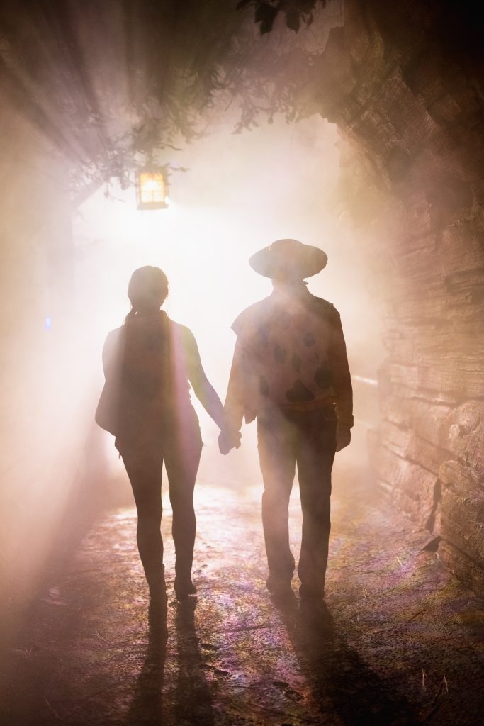 Two guests holding hands in a silhouette picture with fog and white light surrounding them. The guest on the left is shorter than the one on the right who wears a cowboy hat. Guests are walking towards the end of the tunnel towards the light.