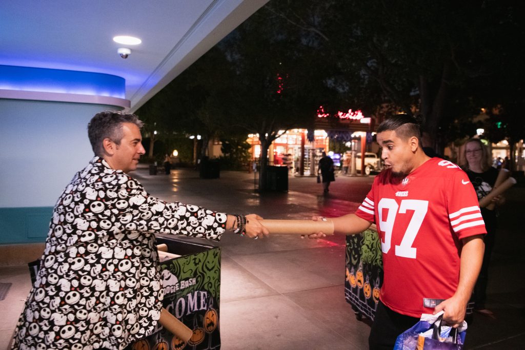 : A cast member handing the commemorative gift, a tubed poster, to a guest leaving the event. The cast member has short dark hair and is wearing a black and white Jack Skellington blazer. The cast member is handing a cardboard tube to the guest. The guest is taking the tube from the cast member. The guest has short dark hair, looks surprised, and is wearing a red and white jersey with the number 97.