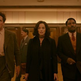 In a still from the film Amsterdam, actors Christian Bale, Margot Robbie, and John David Washington walk side by side down a hallway. Bale wears a brown suit with eggshell button-down dress shirt. His brown tie is cut at his sternum; he holds a brown hat in his right hand. Robbie wears a brown ascot around her neck, with a long black overcoat. Washington wears a black suit, orange vest, red tie, and eggshell button-down dress shirt. He holds a brown hat in his right hand. The lighting is sepia-toned, and a large set of four wooden doors are seen behind them.