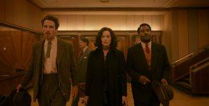 In a still from the film Amsterdam, actors Christian Bale, Margot Robbie, and John David Washington walk side by side down a hallway. Bale wears a brown suit with eggshell button-down dress shirt. His brown tie is cut at his sternum; he holds a brown hat in his right hand. Robbie wears a brown ascot around her neck, with a long black overcoat. Washington wears a black suit, orange vest, red tie, and eggshell button-down dress shirt. He holds a brown hat in his right hand. The lighting is sepia-toned, and a large set of four wooden doors are seen behind them.