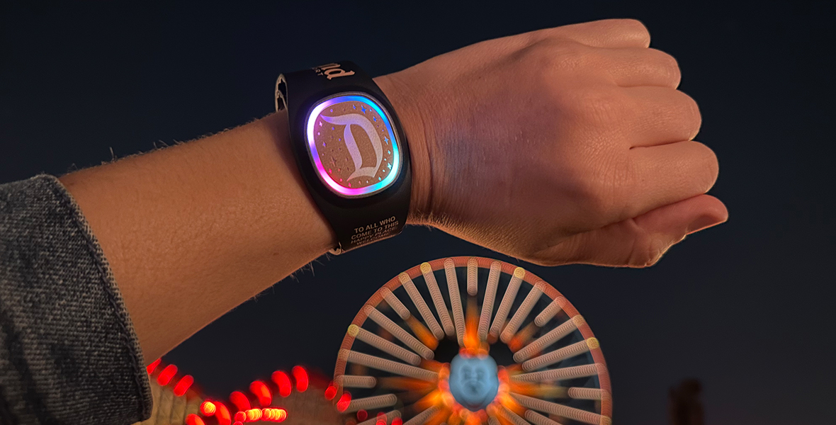 An arm is coming into frame from the left side of the image and a MagicBand+ wristband is on the person’s wrist. The band has a Disneyland “D” on the watch face and it is illuminated in purple and blue tones. Blurry in the background behind the arm is the Pixar Pal-Around Ferris wheel at Disney California Adventure.