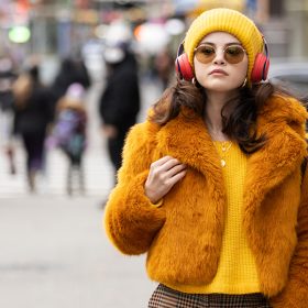 Selena Gomez in a still from Only Murders in the Building. She is wearing yellow plaid pants, a yellow knit sweater, a fluffy orange jacket on top, a yellow beanie, round yellow sunglasses, and red over-the-ear headphones. She is walking down a busy New York City street, but the environment around her is blurred.