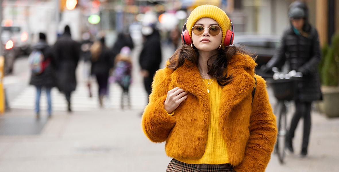Selena Gomez in a still from Only Murders in the Building. She is wearing yellow plaid pants, a yellow knit sweater, a fluffy orange jacket on top, a yellow beanie, round yellow sunglasses, and red over-the-ear headphones. She is walking down a busy New York City street, but the environment around her is blurred.
