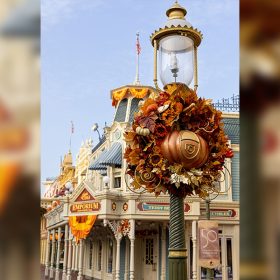 A light post on Main Street, U.S.A., has a large autumnal wreath hanging on it. The wreath is shades of orange and brown. The center of the wreath is a pumpkin.