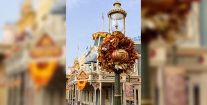 A light post on Main Street, U.S.A., has a large autumnal wreath hanging on it. The wreath is shades of orange and brown. The center of the wreath is a pumpkin.