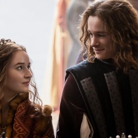 Rosaline (Kaitlyn Dever) stands next to Romeo (Kyle Allen) in an outdoor market at a booth for jewelry, as they look at one another and smile. Rosaline is dressed in a burgundy dress with ornate gold details and a jeweled headband. Romeo wears a dark vest with silver perpendicular details over a long-sleeved shirt, with his shoulder length curly hair framing his face. In the foreground left is Rosaline’s friend and confidante Paris (Spencer Stevenson), who wears a burgundy and gold hat and burgundy velvet shirt with a patterned vest.