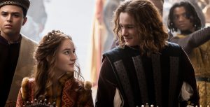 Rosaline (Kaitlyn Dever) stands next to Romeo (Kyle Allen) in an outdoor market at a booth for jewelry, as they look at one another and smile. Rosaline is dressed in a burgundy dress with ornate gold details and a jeweled headband. Romeo wears a dark vest with silver perpendicular details over a long-sleeved shirt, with his shoulder length curly hair framing his face. In the foreground left is Rosaline’s friend and confidante Paris (Spencer Stevenson), who wears a burgundy and gold hat and burgundy velvet shirt with a patterned vest.
