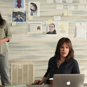 Actor Grace Dove stands with her hands folded at her waist. She holds a black cell phone in her hands. She wears an olive button-down dress shirt with gray pants. Actor Hilary Swank sits in a chair at a desk with a silver laptop turned on in front of her. She wears a black, button-down dress shirt. Across the table is the back of Jeff Perry’s head. He wears a black, button-down dress shirt. On the desk are papers and a white and black coffee mug. On the wall behind Dove are color photos, graphs, and maps pinned to the wall.