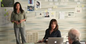 Actor Grace Dove stands with her hands folded at her waist. She holds a black cell phone in her hands. She wears an olive button-down dress shirt with gray pants. Actor Hilary Swank sits in a chair at a desk with a silver laptop turned on in front of her. She wears a black, button-down dress shirt. Across the table is the back of Jeff Perry’s head. He wears a black, button-down dress shirt. On the desk are papers and a white and black coffee mug. On the wall behind Dove are color photos, graphs, and maps pinned to the wall.