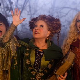 From left to right, Mary, Winifred, and Sarah Sanderson stand next to each other. Mary (Kathy Najimy) has a tall tower of twisted hair atop her head and is wearing a dark maroon cape. Winifred (Bette Midler) has red curly hair loosely shaped like horns. She wears a green dress with a matching cape. Sarah (Sarah Jessica Parker) has long blonde curly hair. She wears a dark red dress, an orange corset, and a dark red cape.
