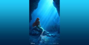 : In the new poster for Disney’s live action The Little Mermaid, Ariel (as portrayed by Halle Bailey) is underwater and sitting pensively on a rock, looking up towards sunlight streaming through the water to her right. Her tail is shades of green, pink, and white, and her long hair is red and in thin braids. Around her are rocks and underwater plant life; beneath it all is sand. On the left at the bottom of the poster is the “Disney” logo, and on the right it says “May 26.”