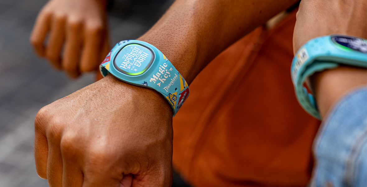 Three hands in fists, each wearing a new MagicBand+ design. The main arm in the center of the image has on a light blue Magic Key holder exclusive MagicBand that reads “The Happiest Place on Earth” across the face.