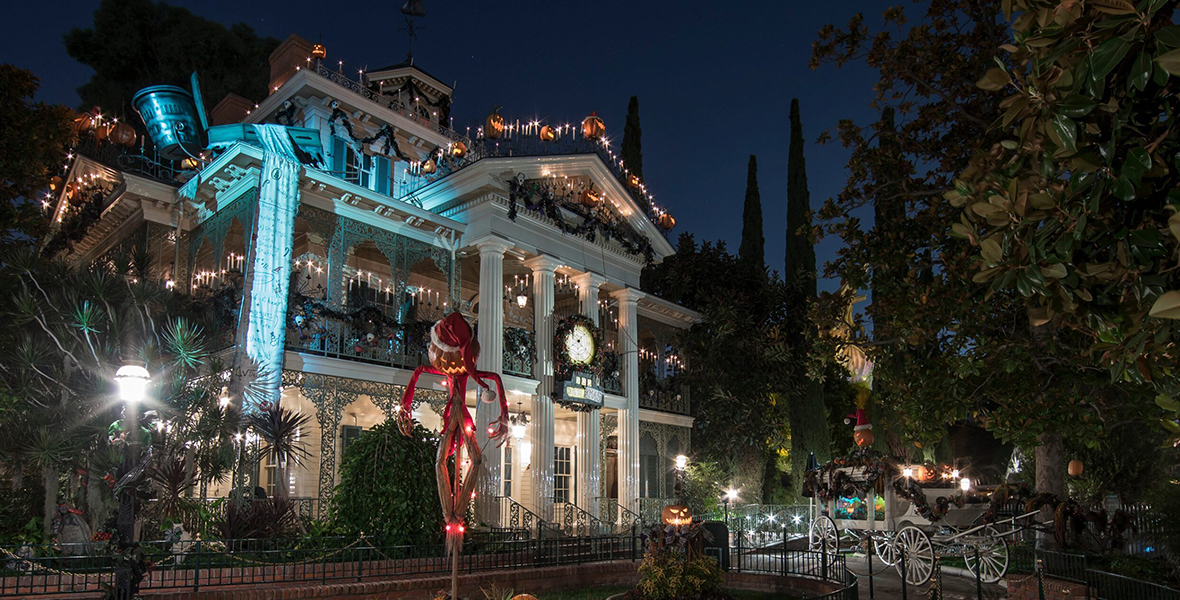 The Haunted Mansion is decorated in Christmas lights and candles. A pumpkin scarecrow stands on the front lawn decorated like Santa Claus. He sits on a bed of pumpkins.