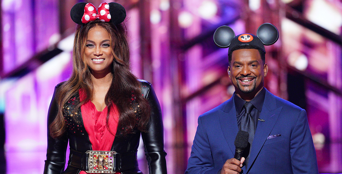Hosts Tyra Banks and Alfonso Ribeiro stand while filming an episode of Dancing with the Stars. Banks wears a Minnie Mouse headband with a red and white polka dot bow. She wears a black leather jacket and red mini dress. She holds a black microphone in her right hand. Ribeiro has black Mickey Mouse ears on top of his head. He wears a blue suit with a dark blue button-down dress shirt.
