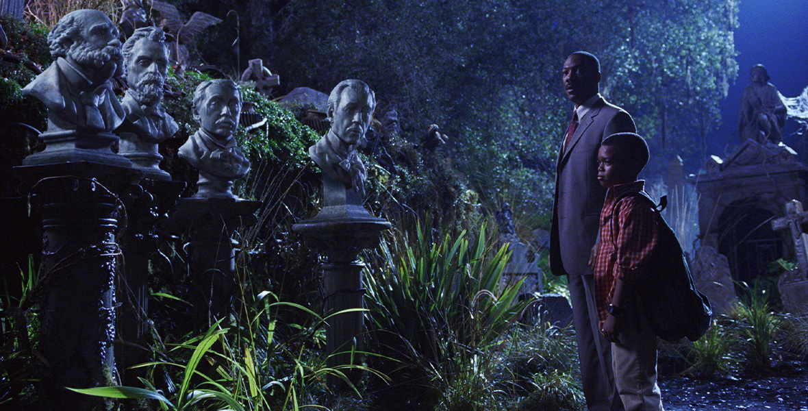 Actors Eddie Murphy and Marc John Jefferies stand side by side in a garden at nighttime. They face four male busts placed atop cement pillars. Murphy wears a gray suit with a white, button-down shirt, and red tie. Jefferies wears a red plaid button-down shirt and tan pants. The garden is full of green-leafed plants and lush trees.