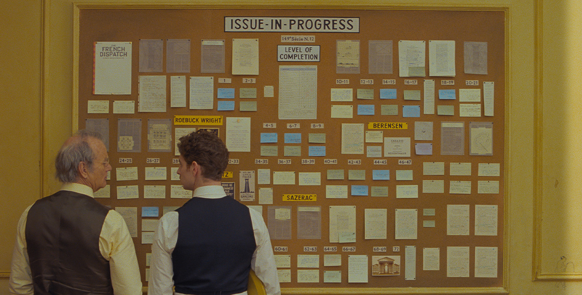 Two men look at each other and face a large bulletin board. The man standing of the left wears a yellow button-down shirt with a brown vest and brown pants. The man standing on the right wears a white button-down shirt with a black vest and black pants. He wears a white apron around his waist and holds a yellow tray in his right arm. The bulletin reads “ISSUE-IN-PROGRESS” at the top with colorful notecards and pieces of paper on the board.