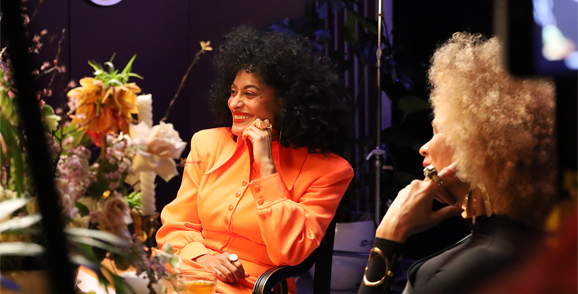 Disney Legend Tracee Ellis Ross sits in a chair with her chin resting on her fist. She smiles and looks on. She wears a bright orange blouse. To the right of the frame is Michaela angela Davis. She wears a black blouse and rests her chin on her fist. The table in front of them has a bouquet of orange and pastel flowers.