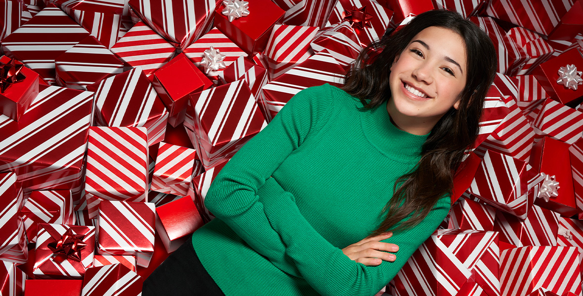 Sarah Nicole Robles is wearing a light-colored sweater and large over-the-ear headphones. She has curly black hair and grins at the camera inside of the recording studio.