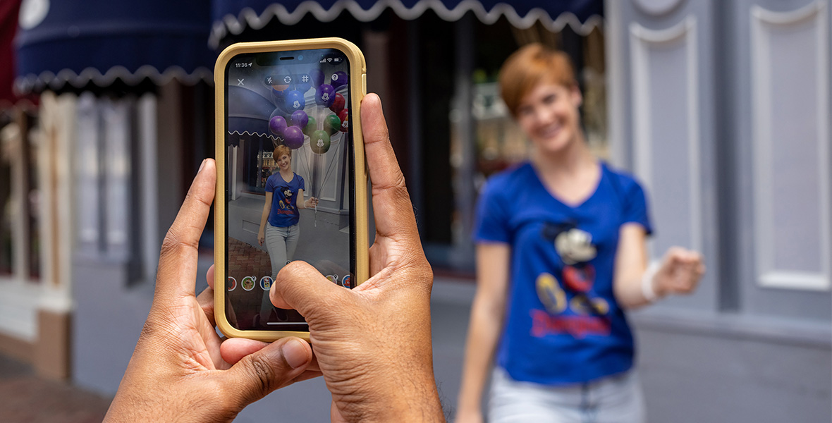In a promotional image for new Disney PhotoPass lenses on the Disneyland app, a woman is having her photo taken on a mobile device (with a yellow-colored case) at Disneyland. The image on the mobile device shows that the PhotoPass lens makes it look like she’s holding a bunch of Mickey Mouse balloons. The woman is wearing a blue Mickey Mouse t-shirt and jeans.