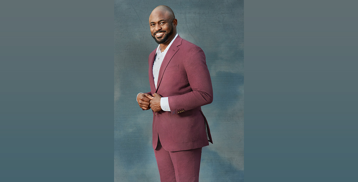 A promo image of Wayne Brady; he is wearing a berry-colored suit with a white button-up shirt, and his hands are at the button of his suit jacket at his waist. He’s standing against a blue-gray background.