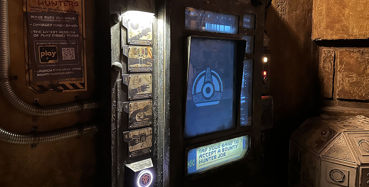 A photo of the Star Wars: Batuu Bounty Hunters mission base inside Star Wars: Galaxy’s Edge. A screen sits in the middle of a highly themed frame made of dirty metal, grates, and signs explaining how to play the game. An illuminated sign below the screen reads, “Tap your band to accept a bounty hunter job.” A glowing circle sits to the left with an illuminated bounty hunter sigil on it—this is where guests tap their MagicBand to receive a mission. 