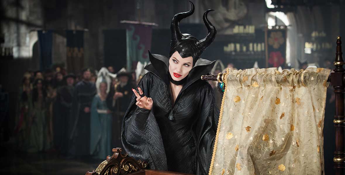 Actor Angelina Jolie portrays Maleficent in the live-action film. She wears a black headpiece with horns, along with a black dress and cape. She leans over a wooden baby crib with gold bedding.