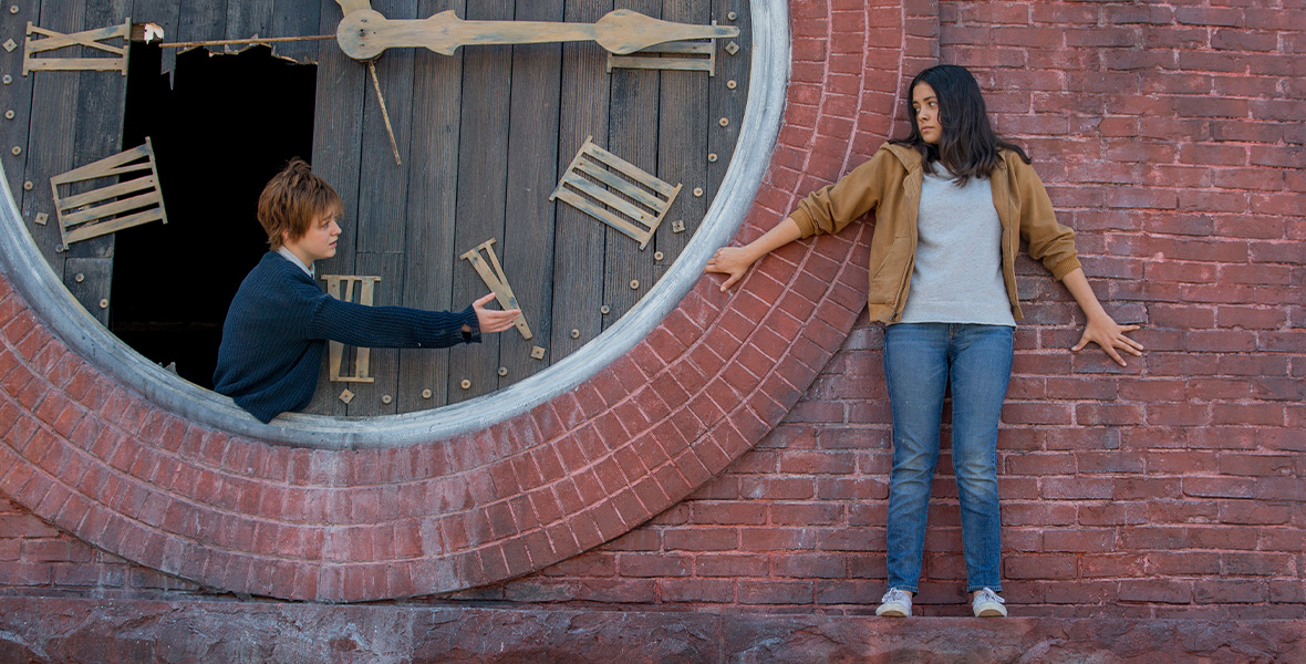 From left to right, actor Maisie Williams leans through a hole in a giant wooden clock on top of a brick building with her arm extended outward. She wears a blue sweater and blue button-down shirt underneath. Actor Blu Hunt leans against the bricks and stands on a ledge. She wears a tan zippered jacket, a gray T-shirt, and blue, denim jeans with white sneakers. Her hands grasp the wall as she looks at Williams.