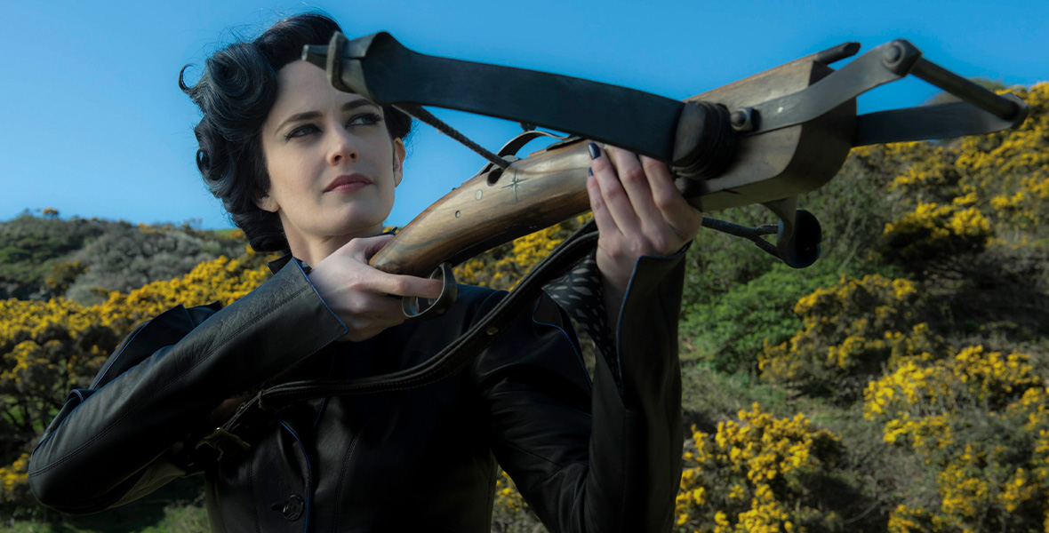 Actor Eva Green portrays Miss Peregrine. She wears a black leather coat and holds a wooden bow and arrow. Behind her is a bright blue sky and lush foliage with yellow flowers.