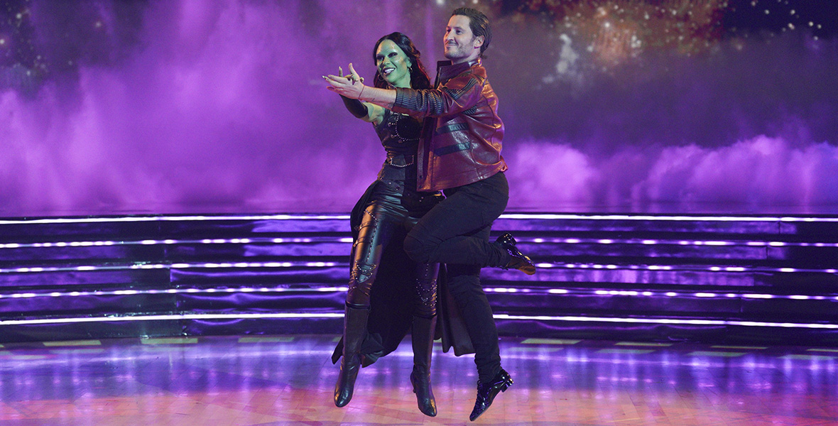 Dancing with the Stars contestants Gabby Windey and Val Chmerkovskiy portray Gamora and Star-Lord from the Guardians of the Galaxy film franchise. Windey wears a black leather ensemble with black leather boots. Windey wears a long black wig and her skin is painted green. Chmerkovskiy wears a maroon leather jacket with black accents, black pants, and black shoes. The screen behind them projects a galactic graphic with purple lights highlighting the stage and dance floor.