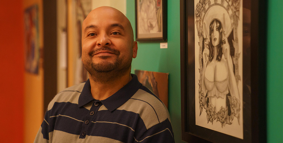 Frankie Quinones is wearing a gray and black striped polo with four buttons. He is bald with a little facial hair. He stands against a green wall with art.