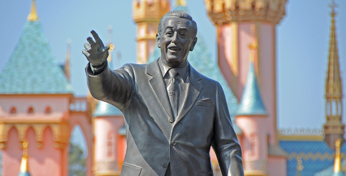 A close-up of Walt Disney as he’s seen in the famous “Partners” statue at Disneyland Park. His arm is raised and he’s looking into the distance. Sleeping Beauty Castle can be seen in the background.