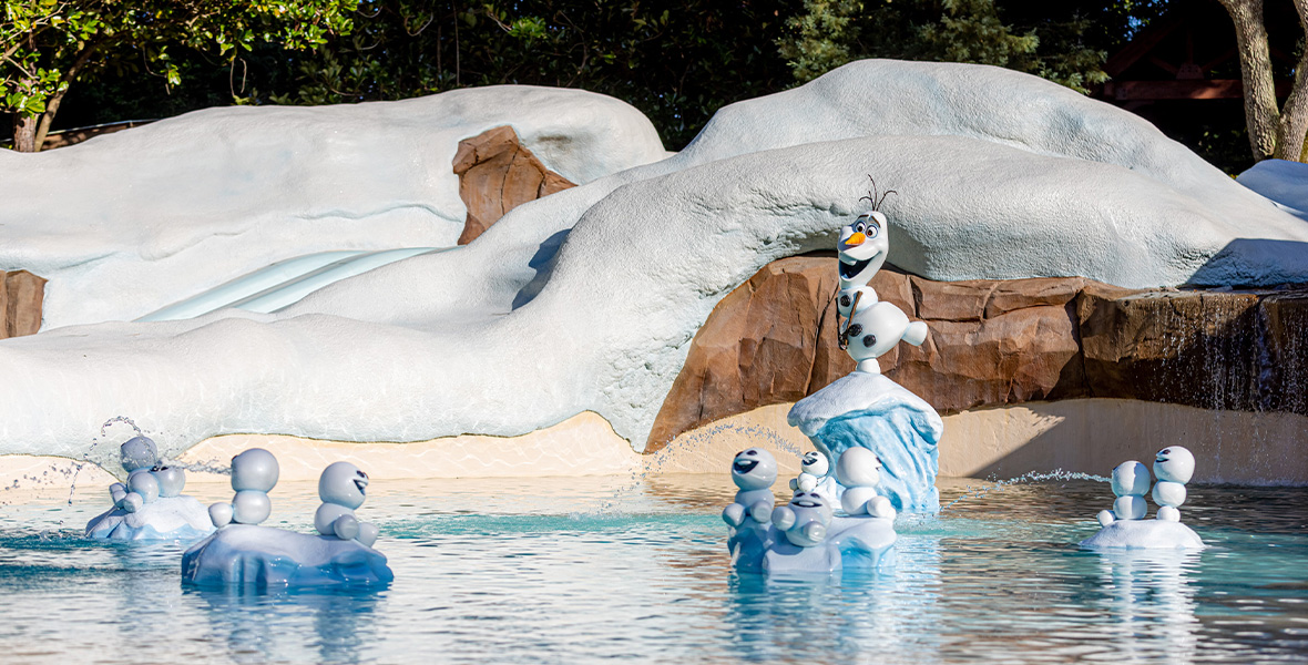 In an image of Blizzard Beach Water Park at Walt Disney World Resort, a small figure of Olaf and figures of his Snowgie pals are seen standing on “snowy” rocks throughout a pool—and a “snow”-covered water slide is seen in the background, as well as some trees. A small waterfall is emptying over some rocks into the pool, to the right.