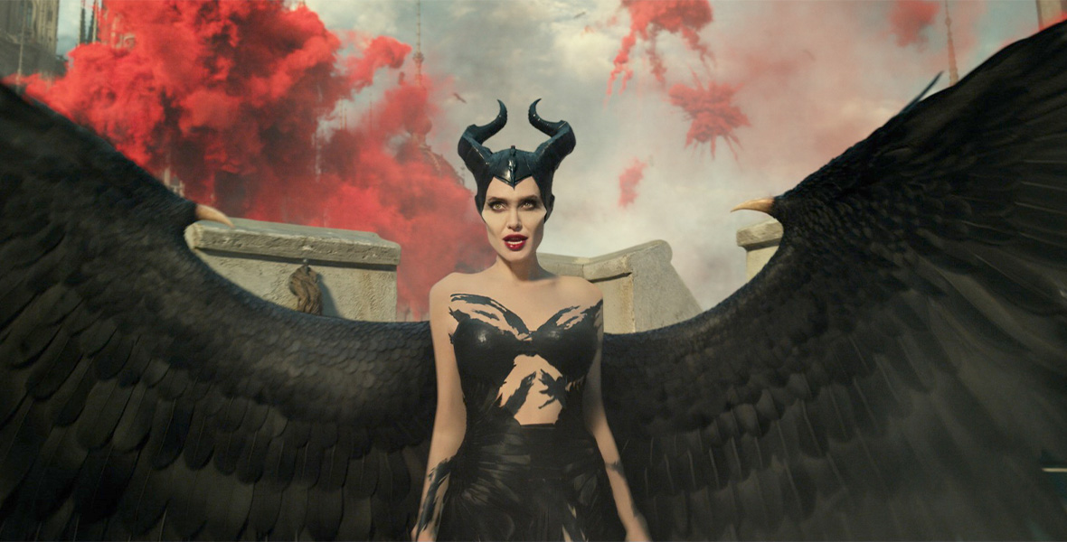 Actor Angelina Jolie portrays Maleficent with a long, black sleeveless gown and black, horned headpiece. Attached to her body are enormous bird-like wings with two sharp horns on each side. Behind her are large, red smoke clouds in the sky.