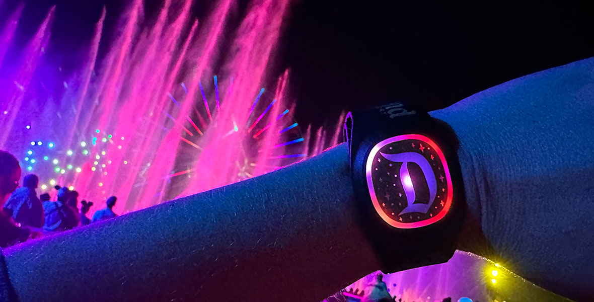 An arm stretched in front of the camera with an illuminated MagicBand+ on the wrist. The band is glowing pink and fountains from Disney California Adventure’s World of Color show are shooting into the air behind the arm.