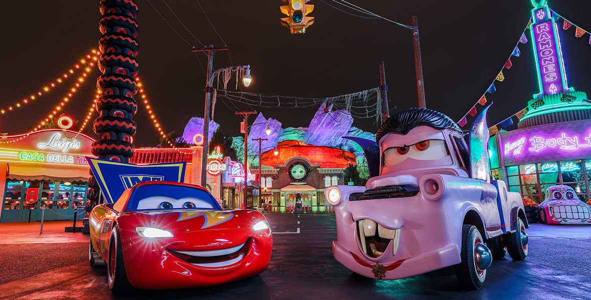Lightning McQueen is a red sportscar. He is dressed up in a blue superhero costume. Mater is to his left. Mater is a purple tow truck and sports a black wig, mustache and vampire fangs.