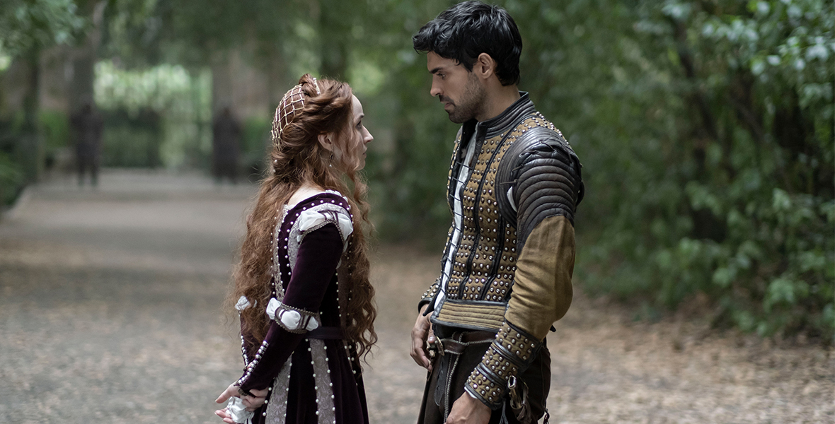 Rosaline (Kaitlyn Dever) and Dario (Sean Teale) stand on a forest path looking at one another. Rosaline wears a burgundy and lace gown with pearl accents, with her hands clasped behind her back—one injured hand is wrapped in muslin. Dario has dark short hair and a light beard. He is wearing a black and tan leather and cloth coat; metal details run up and down the front and forearms of the garment, a sword visible in a holster at his hip.