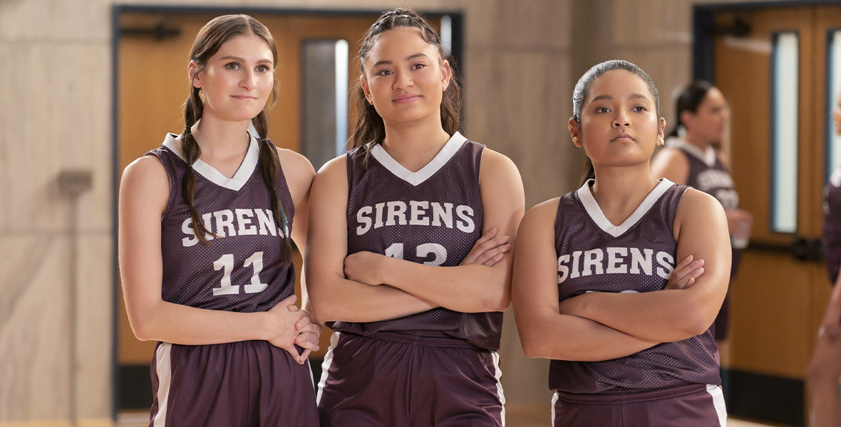 From left to right, actors Cricket Wampler, Tiana Le, and Tisha Custodio stand side by side and fold their arms. They wear dark purple jerseys with white piping and “Sirens” spelled across the front and their respective number. The shorts have a logo on the lower right side.