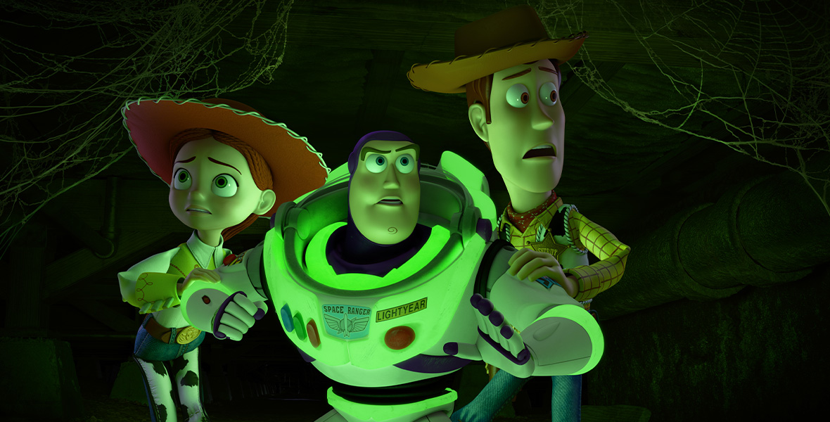 From left to right, animated characters Jessie, Buzz Lightyear, and Woody stand hunched and huddled together. They are wide-eyed and appear scared. Jessie wears a red cowboy hat with a white shirt, a yellow vest, and jeans with cow-print chaps. Buzz stands with his arms extended and wears a white space suit. Woody wears blue jeans, a brown cowboy hat, and a yellow and red button-down shirt. Cobwebs are seen above them in each corner of the frame.