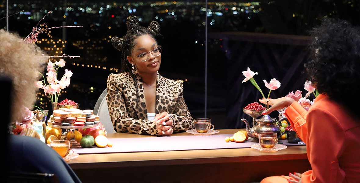 Actor Marsai Martin sits at a large wooden table with her hands folded. She wears a leopard print suit. The desk has vases with orange, yellow, and pink flowers, and knickknacks. Opposite Martin sits Michaela angela Davis and Disney Legend Tracee Ellis Ross. Ross wears an orange blouse while Davis wears a jewel-toned blue blouse.