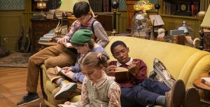 Actors Mystic Inscho, Emmy DeOliveira, Marta Kessler, and Seth B. Carr sit on a mustard couch and each hold open books. Behind them are tables and furniture with stacks of books and knickknacks. A red bucket sits at the edge of the couch in front of DeOliveira.
