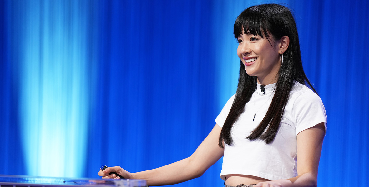 Actor Constance Wu smiles and stands at a large podium with a glass top. She wears a white, cropped T-shirt and tan pants with a black pattern. She holds a buzzer in her right hand. Behind her is a bright blue backdrop.