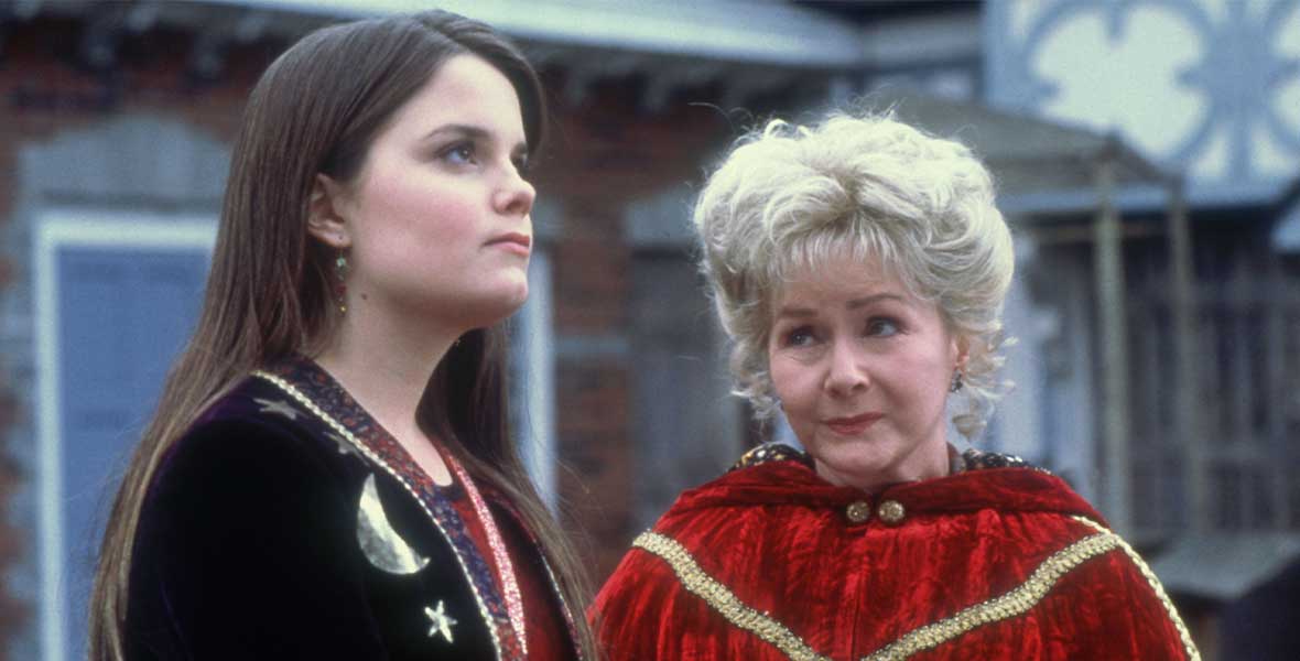 In a still from Halloweentown II: Kalabar’s Revenge, actor Kimberley J. Brown wears a long black cloak with embroidered yellow stars. She looks up as actor Debbie Reynolds stands to her left; Reynolds wears a red velvet cloak with gold stitching.