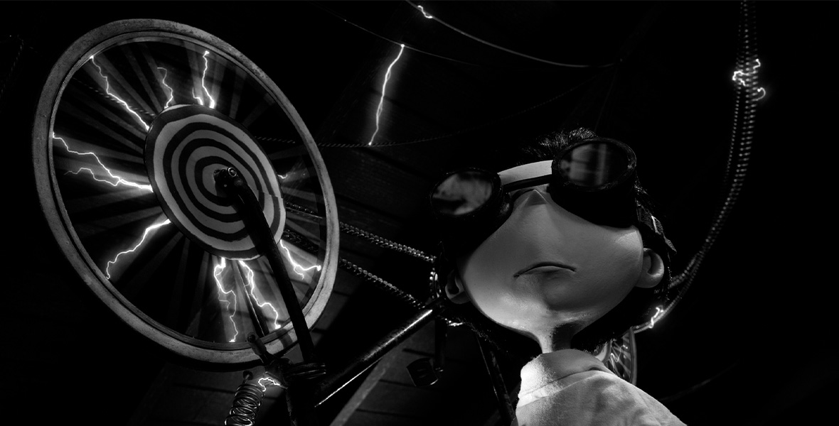 An animated young boy wears a white coat and large black goggles. He stands next to a spinning wheel that has lightning spurting out and black and white accents.